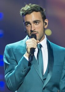 Marco Mengoni im blauen Anzug singt in ein handmikrofon und blickt in Richtung Kamera