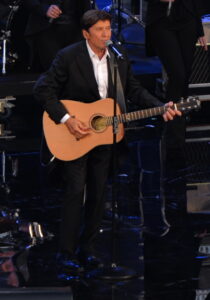 Gianni Morandi steht 2016 mit Gitarre vor einem Ständermikrofon auf der Bühne der Arena von Verona