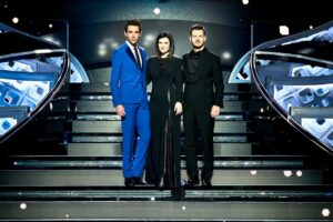 Mika, Laura Pausini und Alessandro Cattelan stehen eng nebeneinander auf den Stufen der Bühne des Ariston-Theaters. Mika trägt einen blauen Anzug, Pausini und Cattelan sind in Schwarz gekleidet.