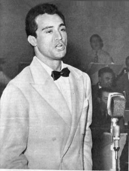 Schwarz-weiß-Foto von Nunzio Gallo in Anzug mit Fliege vor einem Standmikrofon, im Hintergrund ist Publikum zu sehen.