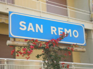 Blaues Schild mit der weißen Aufschrift SAN REMO vor einem Gebäude, geziert von roten Blumen