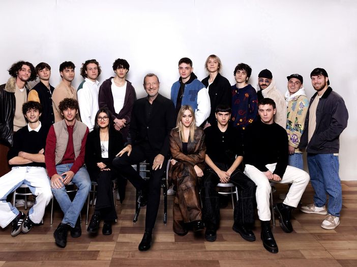 Die Teilnehmenden von Sanremo Giovani 2022 vor weißem Hintergrund in zwei Reihen, die hintere Reihe stehend, die vordere Reihe auf Stühlen sitzend. In der Mitte sitzt Amadeus auf einem Stuhl.