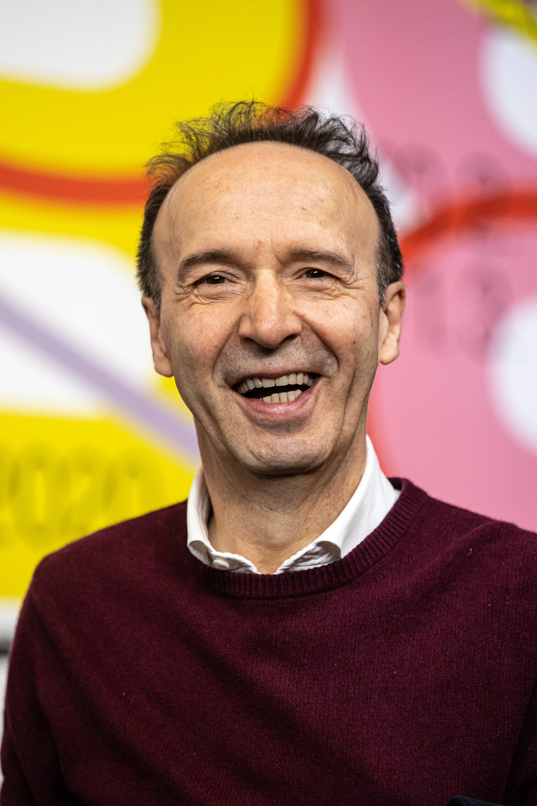 Frontalaufnahme von Roberto Benigni. Er lacht in die Kamera und trägt einen roten Pullover.