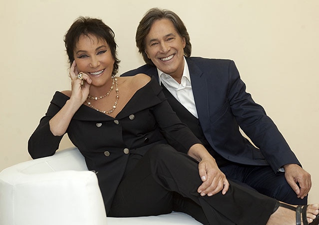 Angela und Angelo von Ricchi e Poveri sitzen nebeneinander auf einem weißen Sofa und tragen dunkle Abendkleidung.