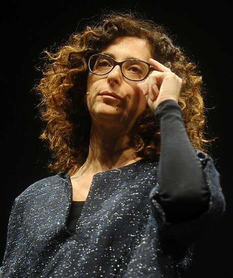 Teresa Mannino vor schwarzem Hintergrund fasst sich mit der linken Hand an ihre Brille. Sie trägt ein schwarzes, weiß gesprenkeltes Oberteil.
