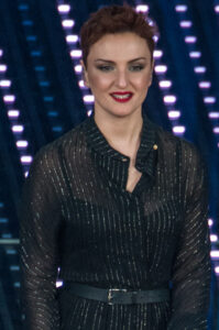 Die Sängerin Arisa steht vor einem beleuchteten Bühnenhintergrund. Sie trägt ein dunkles Kleid mit offenem Kragen.