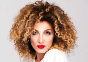 Nahaufnahme von Marcella Bella auf Schulterhöhe. Sie trägt ein weißes Oberteil und blickt direkt in die Kamera