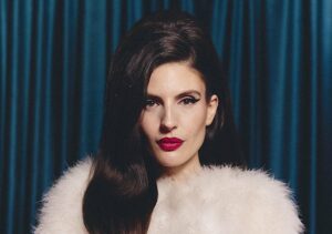 Poträtfoto von Ditonellapiaga vor einem dunkelgrünen Vorhang. Sie trägt ein weißes Pelzoberteil.