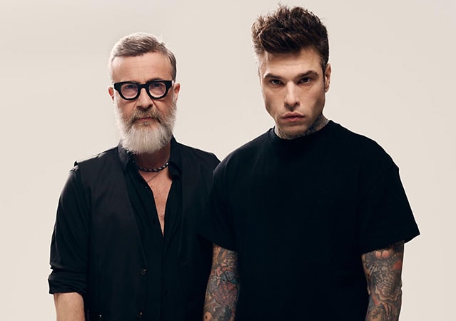 Marco Masini und Fedez stehen vor neutralem Hintergrund, die untere Körperhälfte ist abgeschnitten. Beide sind in Schwarz gekleidet und blicken ernst in die Kamera.