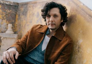 Ermal Meta sitzt an die Wand gelehnt in einem Treppenhaus und blickt freundlich in die Kamera. Er trägt eine hellbraune Jacke, ein türkises, ausgeschnittenes Oberteil und ein weißes Unterhemd.