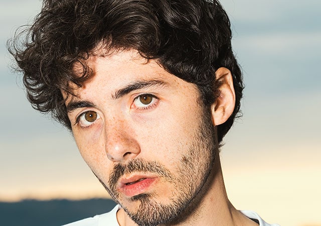 Fulminacci mit dunklem, leicht gewelltem Haar vor einem hellen Hintergrund. Er blickt ruhig und direkt in die Kamera.