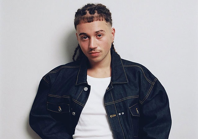 Sayf mit eng geflochtenen Haaren, getragenem Jeansjackett mit gelben Nähten und weißem Shirt, vor hellem Hintergrund stehend und mit neutralem Blick in die Kamera.