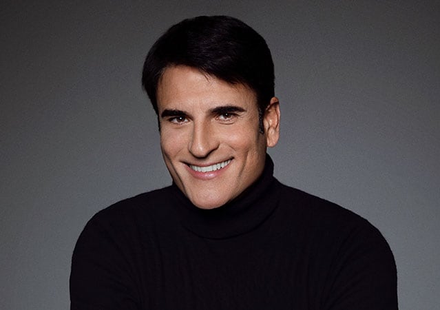 Porträt Sal Da Vincis mit kurzem dunklem Haar und schwarzem Rollkragenpullover vor dunklem Hintergrund, freundlich lächelnd und direkt in die Kamera blickend.