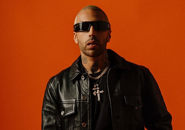 Luchè mit blond gefärbtem Kurzhaarschnitt, schwarzer Lederjacke und dunkler Sonnenbrille vor kräftig orangefarbenem Hintergrund; er trägt eine auffällige Kette mit Kreuzanhänger und zeigt ein neutrales Gesicht.