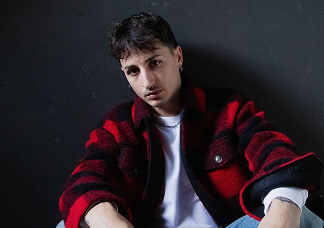 Nayt sitzt vor dunklem Hintergrund, trägt eine rot‑schwarze Karojacke über einem weißen Shirt und hellblaue Jeans. Er hat kurzes dunkles Haar, Ohrringe und eine Kette und blickt mit neutralem Ausdruck direkt in die Kamera.