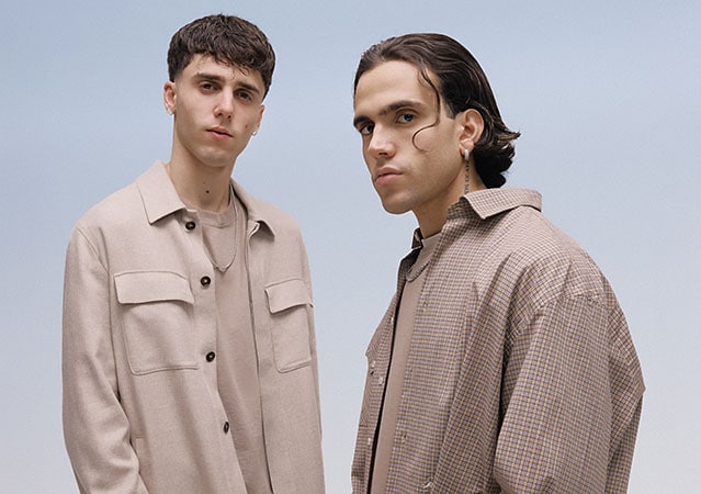 LDA und Aka 7even stehen nebeneinander vor einem neutralen Hintergrund. Beide blicken direkt in die Kamera. Das Porträt wirkt professionell ausgeleuchtet und zeigt die beiden Künstler in halbnaher Einstellung.