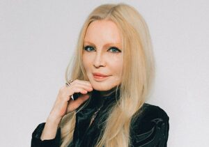 Porträt von Patty Pravo, frontal aufgenommen, mit warmem Bühnenlicht und ruhigem Gesichtsausdruck. Sie hat langes, blondes Haar, und trägt ein dunkles Oberteil.