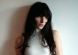 Angelica Bove mit langen dunklen Haaren steht vor einem hellen, schlichten Hintergrund. Sie trägt ein ärmelloses weißes Rollkragen‑Top und blickt leicht zur Seite, mit einem ruhigen, nachdenklichen Ausdruck.