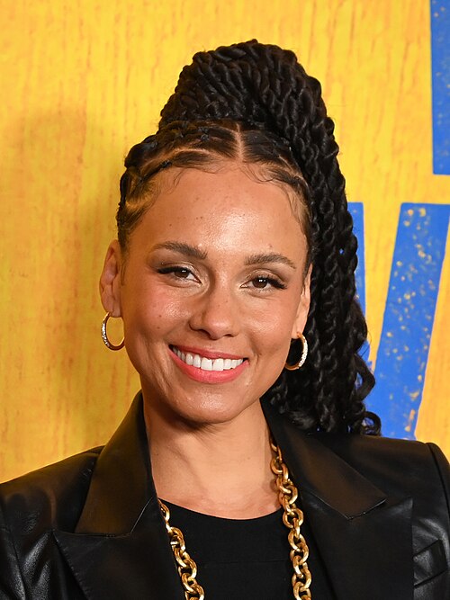 Porträt von Alicia Keys, aufgenommen im April 2025 in Manhattan. Sie steht lächelnd vor der Kamera, trägt eine schwarze Lederjacke, goldene Ketten und lange geflochtene Haare. Ihr Gesicht ist frontal zu sehen, die Beleuchtung ist weich, und der Hintergrund bleibt neutral, sodass der Fokus klar auf ihr liegt.