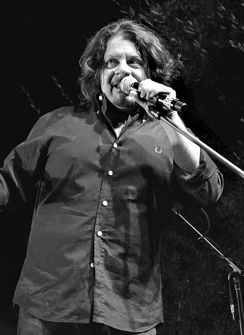 Schwarzweißfoto von Lillo im schwarzen Hemd, der vor einem Standmikrofon steht; er umklammert das Mikrofon mit der linken Hand