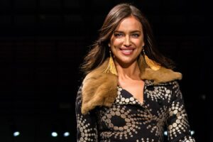 Irina Shayk vor dunklem Hintergrund, sie trägt ihr Haar offen, lange Ohrbehänge und ein gemustertes Oberteil mit einer Pelzkrempe