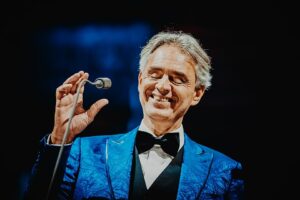 Andrea Bocelli während eines Konzerts im Stadion von Poznań im Mai 2019. Er steht auf der Bühne, elegant gekleidet, und singt in ein Mikrofon. Die Beleuchtung ist warm und hebt sein Gesicht sowie die Konturen seines blauen Anzugs hervor. Der Hintergrund bleibt dunkel, wodurch der Fokus klar auf ihm als Performer liegt.