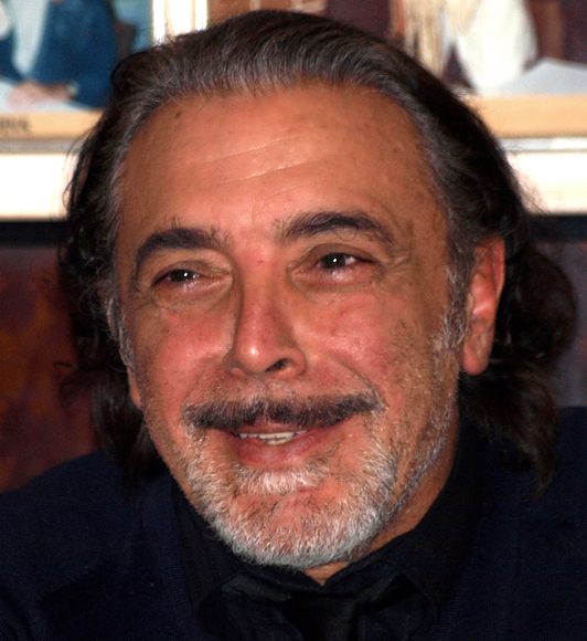 Porträt des italienischen Schauspielers Nino Frassica, aufgenommen 2012: Er blickt leicht zur Seite, trägt ein dunkles Jackett und steht oder sitzt vor neutralem Hintergrund.