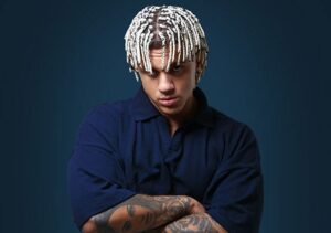 Samurai Jay in blauem Hemd und mit auffälligen, blonden Drills hält den Kopf leicht gesenkt und die Arme verschränkt. Der Hintergrund ist neutral und auf seinen Armen sind Tätowierungen zu erkennen.