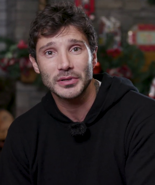 Porträt des italienischen TV‑Moderators Stefano De Martino: Er steht vor einem neutralen Hintergrund, trägt einen dunklen Pullover und blickt leicht lächelnd in die Kamera.