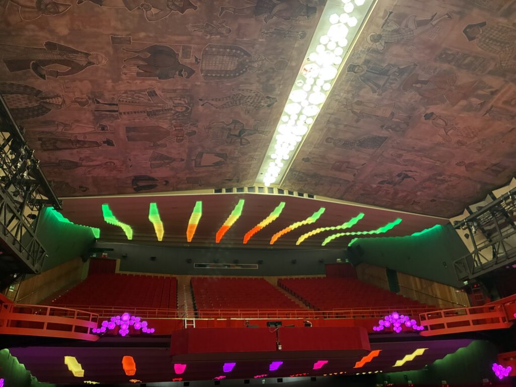 Blick in die Galerie des Ariston-Theaters in Sanremo: rote Sitzreihen und Balkone mit leuchtenden grünen Akzenten, darüber eine kunstvoll bemalte Decke mit Figurenmotiven. Das Licht taucht den Saal in warme, lebendige Farben und betont die elegante Architektur des traditionsreichen Hauses.