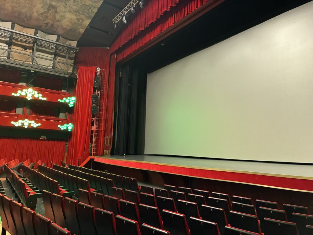 Großer Saal des Ariston-Theaters in Sanremo: Blick auf die Bühne mit geöffnetem roten Vorhang und weißer Kinoleinwand, umgeben von leuchtend roten Wänden und Sitzreihen. Links sind mehrere Ränge mit roten Paneelen und grünen Lichtakzenten zu sehen, darüber die kunstvoll bemalte Decke. Die Perspektive zeigt die elegante, klassische Atmosphäre des berühmten Veranstaltungsorte.