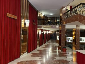 Eingangsbereich des Ariston-Theaters in Sanremo: ein großzügiges Foyer mit roten Samtvorhängen, glänzendem Marmorboden im geometrischen Muster, hohen Säulen und funkelnden Kronleuchtern. Entlang der Wände stehen beleuchtete Vitrinen, darüber Wegweiser zu den Sitzbereichen II settore und Poltronissime. Eine Galerie mit verziertem Geländer bildet den oberen Abschluss des Raums.